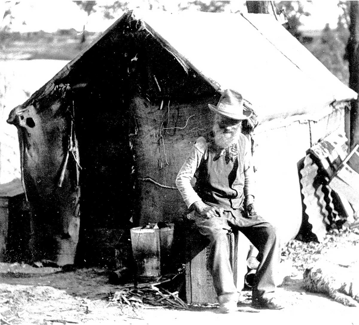 Old man seated on a crate in front of his simple dwelling with a dog at his feet.