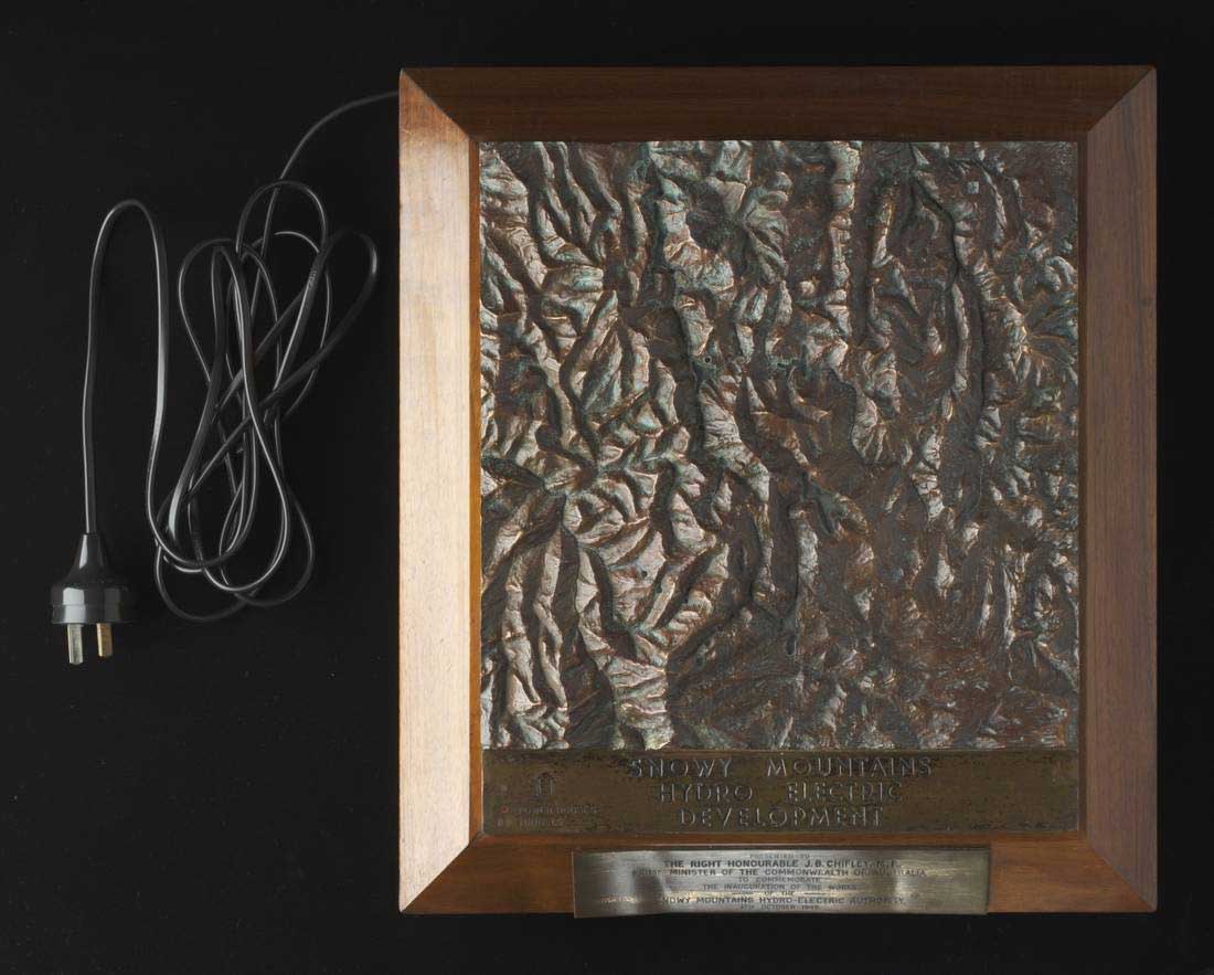 A bronze topographic model of the Snowy Mountains Hydro Electric Scheme, mounted on a brown wooden plinth with four black rubber feet.