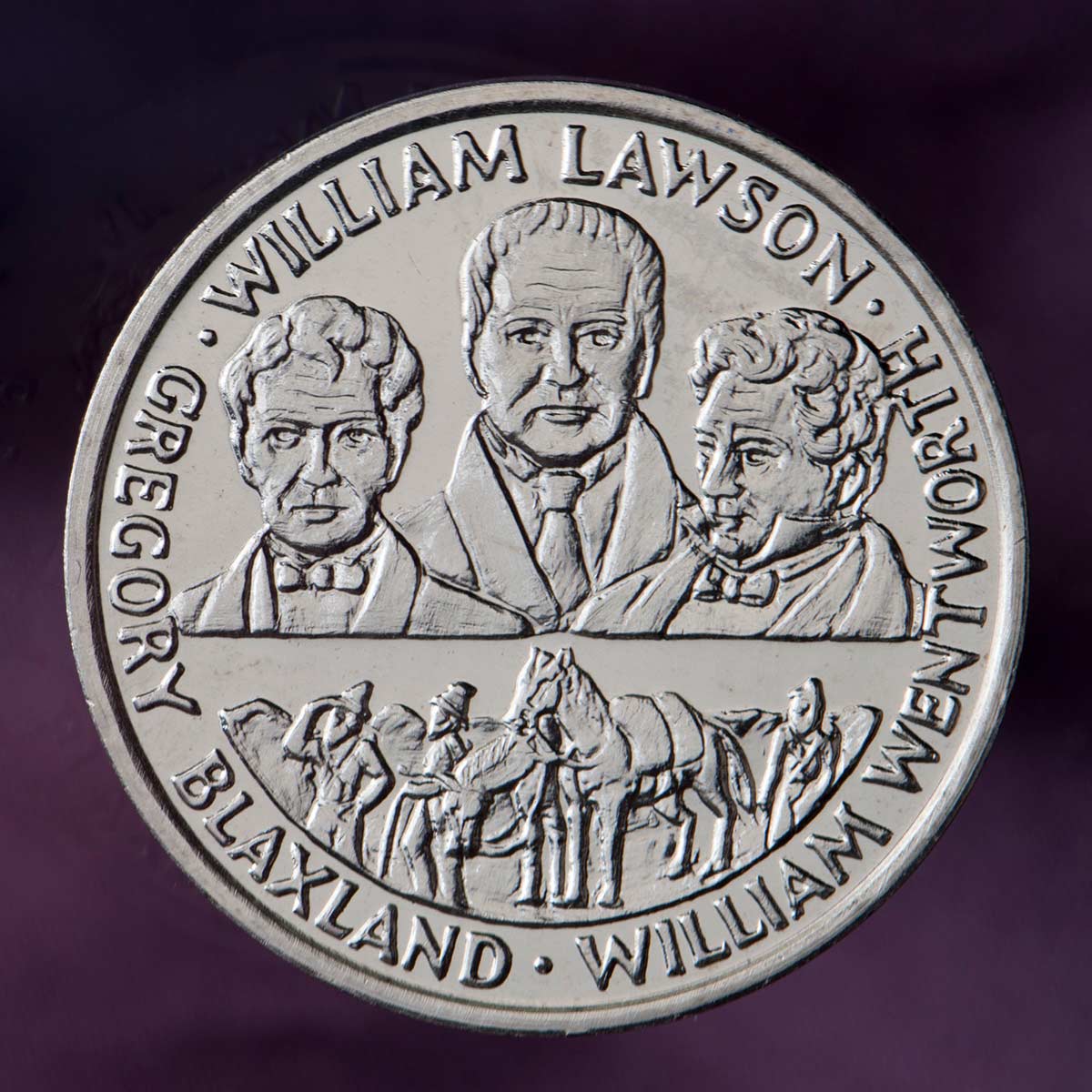 A silver medal against a purple background. In the top half are portraits of Gregory Blaxland, William Wentworth and William Lawson, and in the bottom half are the men with horses against a mountain backdrop.