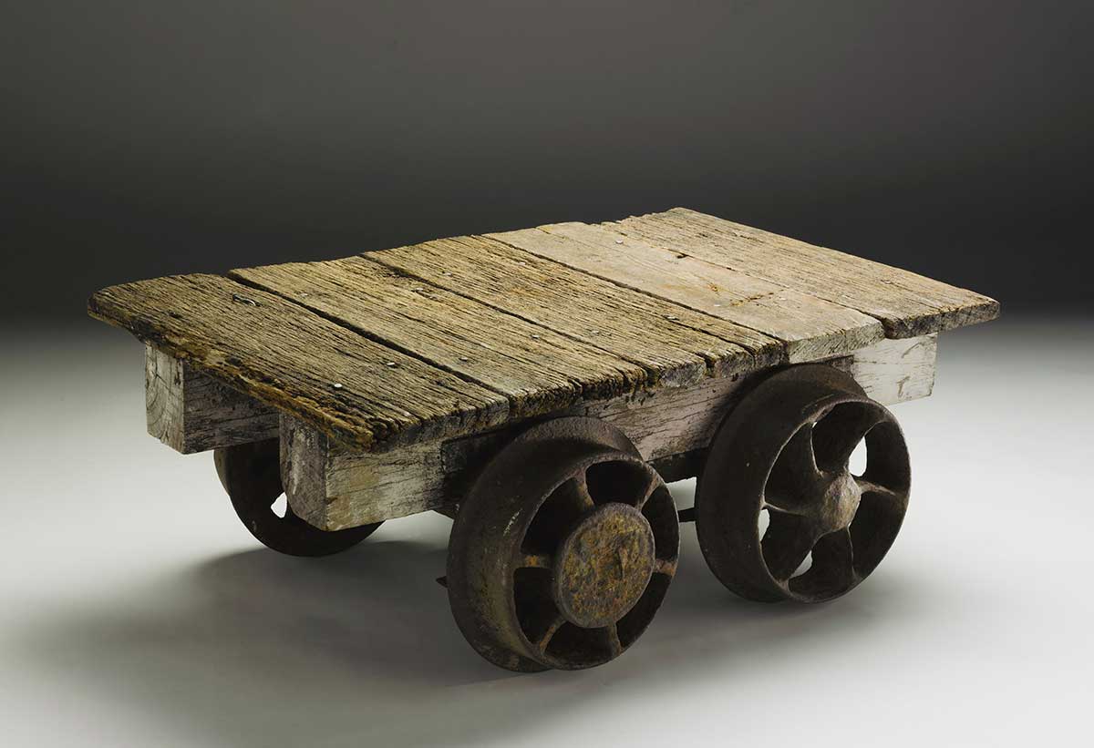 A flat topped brown coloured wooden cart with four rusted metal wheels. One of the planks of wood of the cart has been replaced and is of a lighter colour.