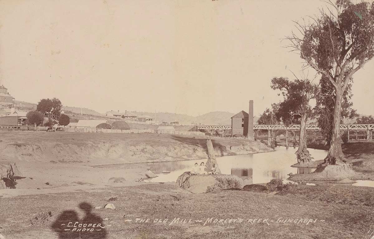 Old black and white photo of the mill in the distance surrounded by bare landscape and a few trees to one side. A river is in the foreground.