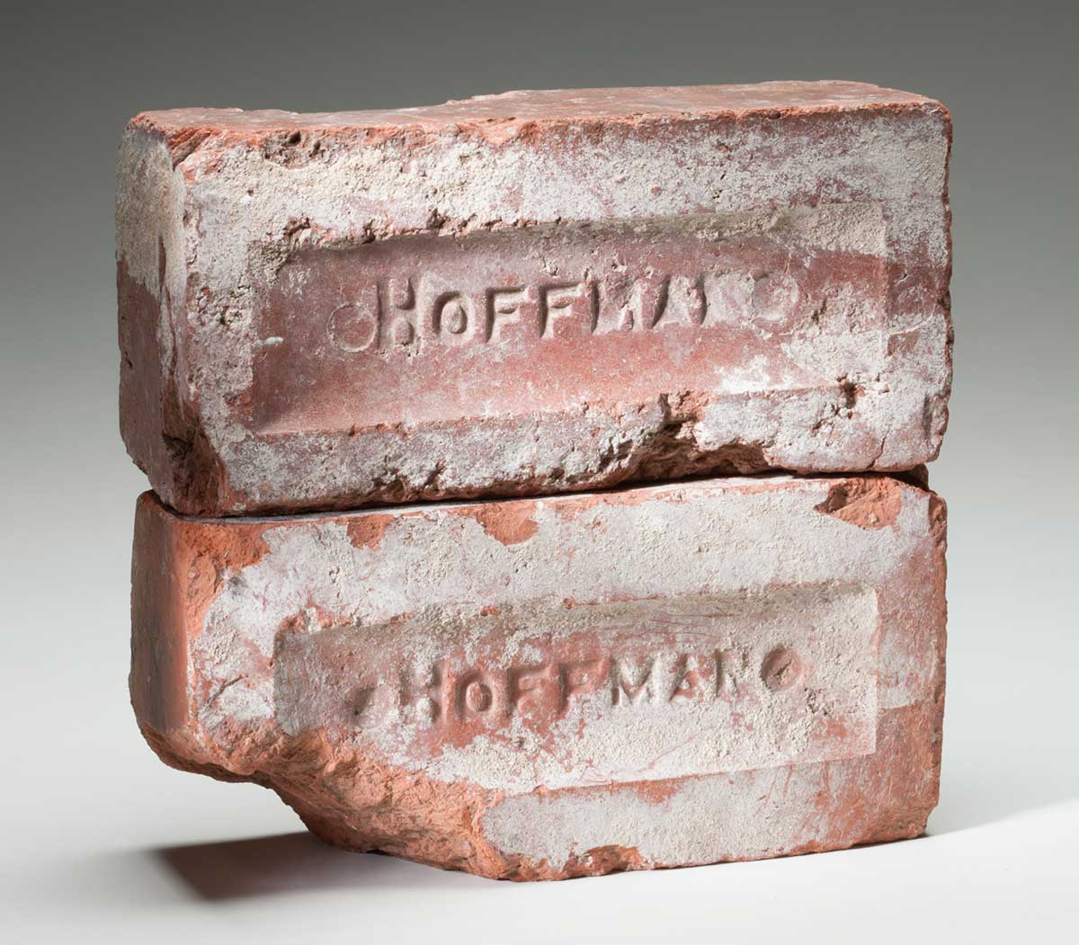 A rectangular red clay and concrete brick. White concrete is coated across four sides of the brick. The word 'HOFFMAN', is imprinted on the top of the brick.