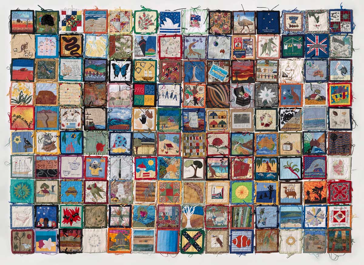 A rectangular patchwork quilt with two separate pieces of black cotton fabric. The quilt is made up of 140 individually designed and sewn square blocks attached together using ribbon ties. The proper top right corner block depicts a small shack, red earth and a blue sky. The quilt blocks attach to a black cloth backing and there is an additional piece of black cotton fabric with raw edges.