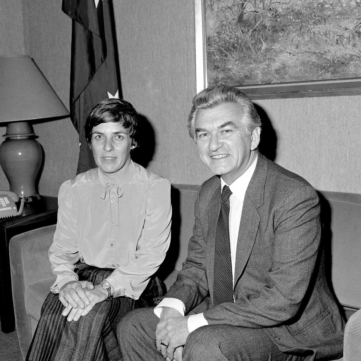 Pam O’Neill and Bob Hawke pose for the camera, seated on a couch.