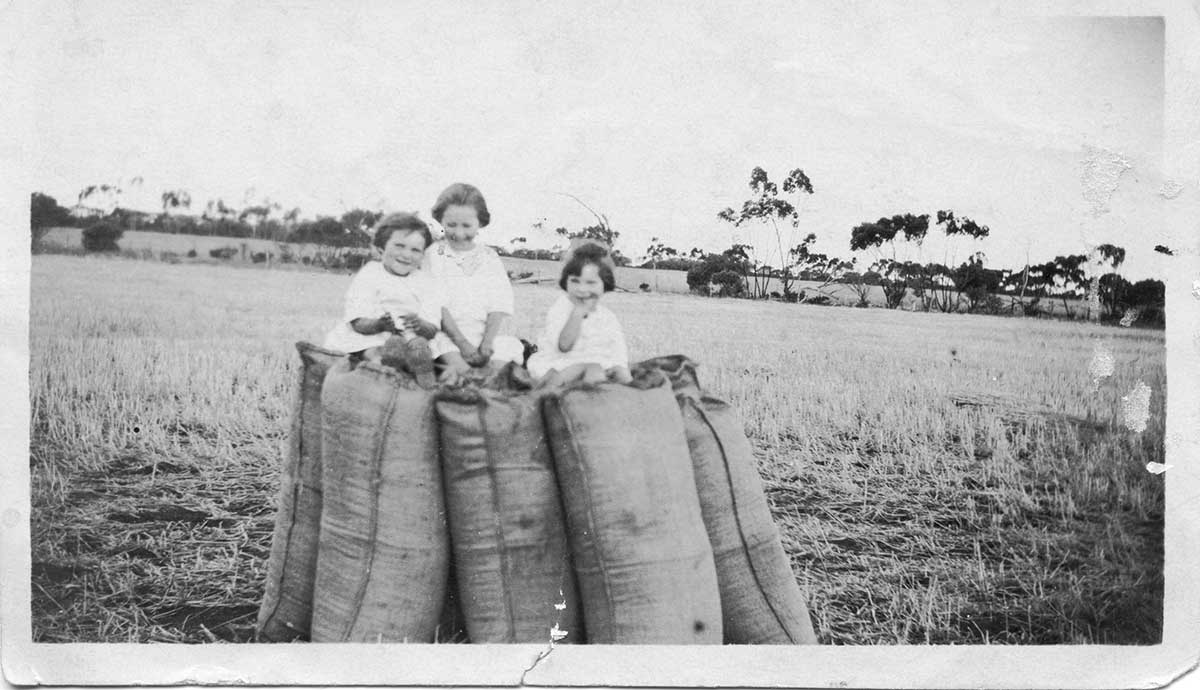 Black and white photograph of three young girls sitting on top of large sacks in paddock.