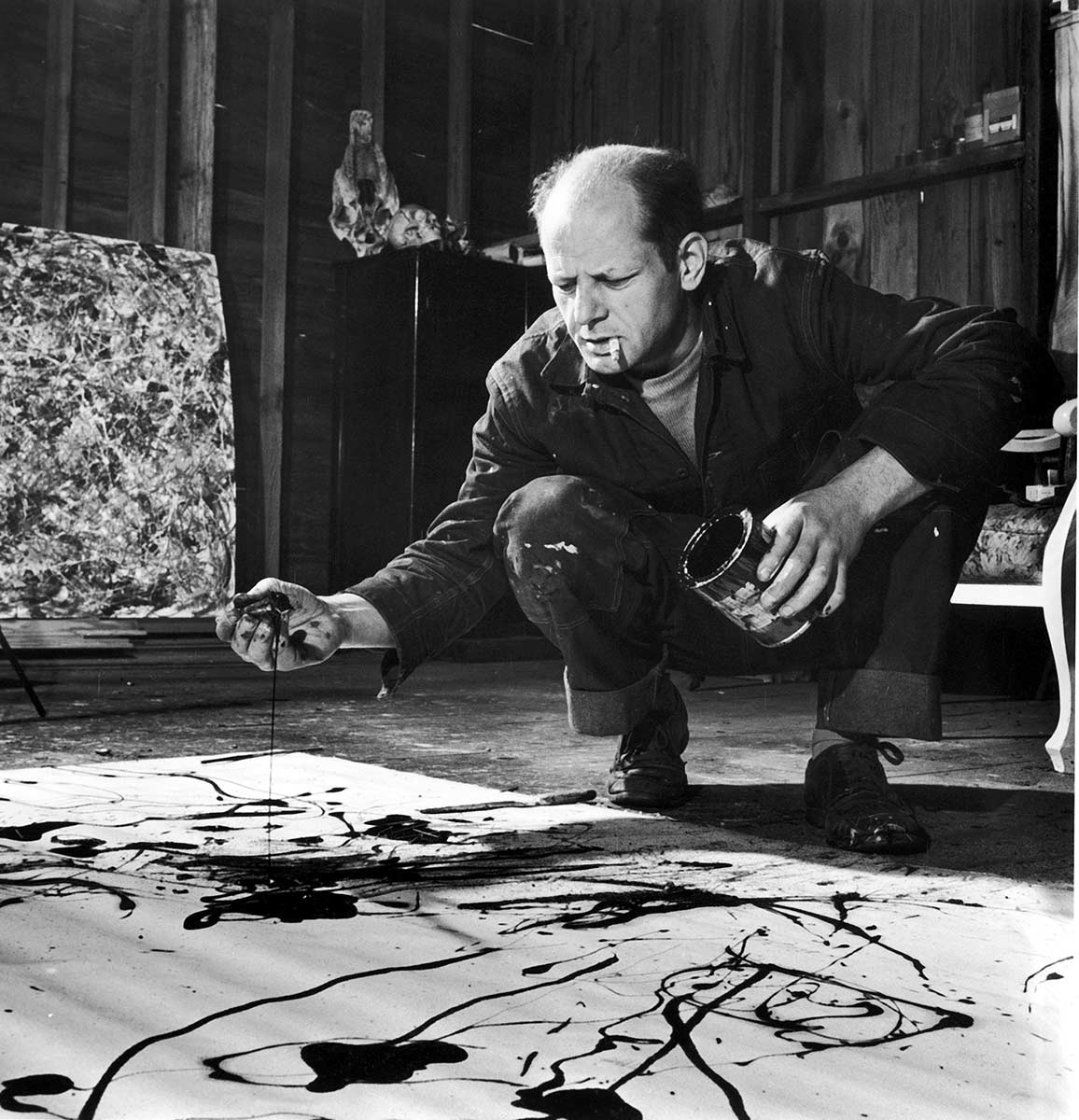 Black and white photo of balding, middle-aged man crouches by canvas spread on the floor. He has a cigarette in his mouth, a paint can in one hand. His other hand seems to be covered in paint. He is frowning intently at the canvas, which has a few splotches of dark paint on it.