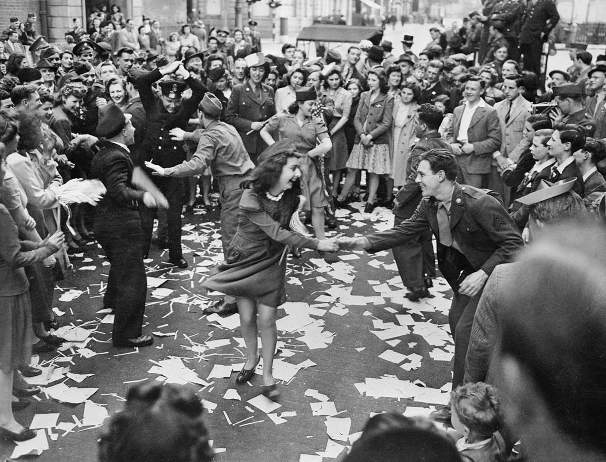 Black and white photograph of a large crowd of citizens celebrating in the street. In the centre of the photograph people are dancing joyously.