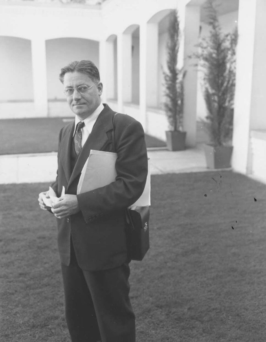 Black and white photograph of a man in a suit and spectacles standing in a courtyard.