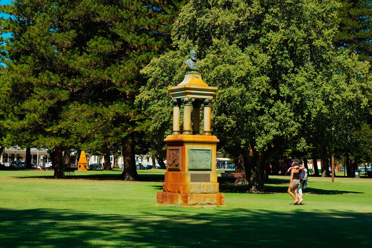 Colour photograph of a memorial with the bust of a man erected at the top. It is situated in a well-maintained public park with large established trees, and a couple leisurely strolling past.