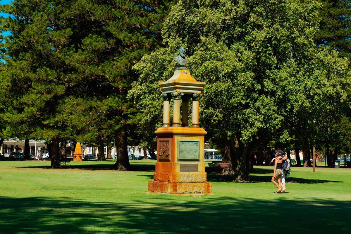 Colour photograph of a memorial with the bust of a man erected at the top. It is situated in a well-maintained public park with large established trees, and a couple leisurely strolling past.