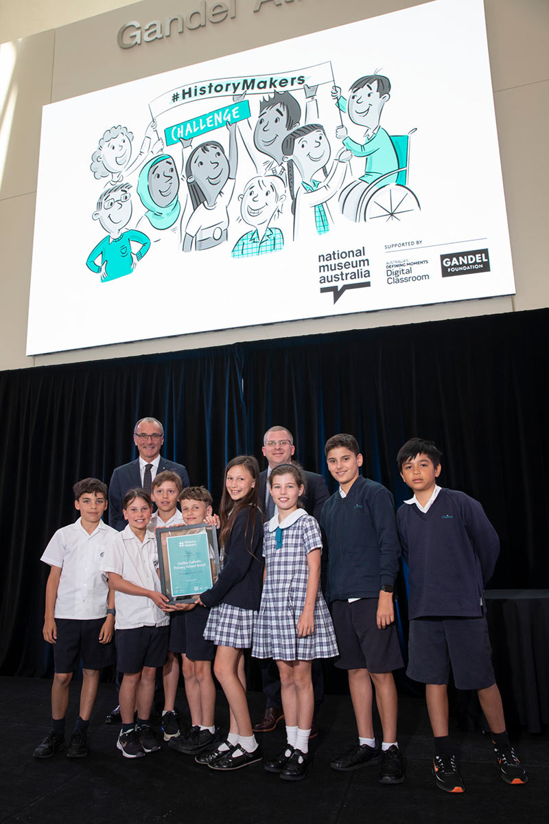 A group of school children and two men are standing on a stage below a digital screen. The screen features a sketch of children with a banner with the text 'History Makers Challenge'.