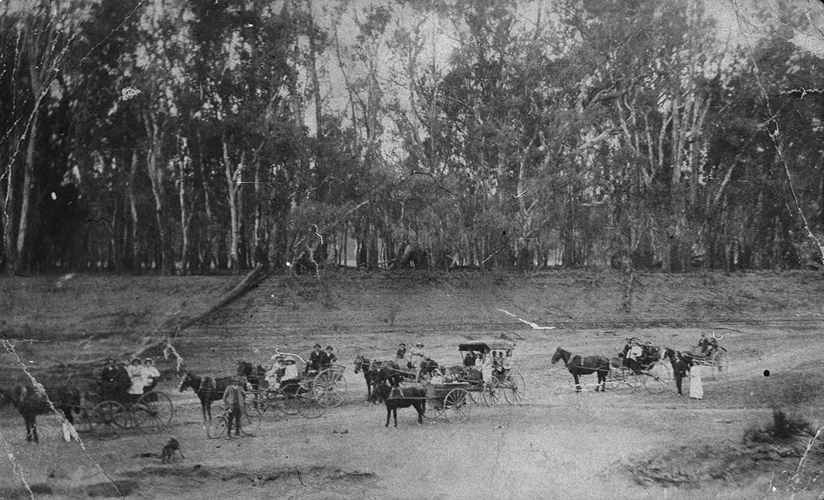 Black and white photograph of a tree-lined dry river bed on which people are standing or seated in horse and carts.