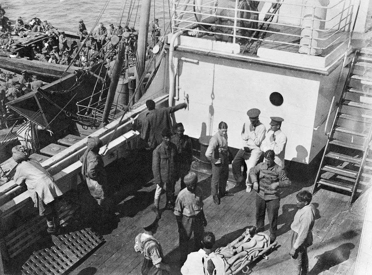 Black and white photograph of men on the deck of a ship with barges containing large groups of men docked below.