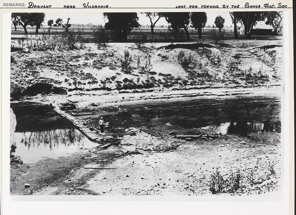 Black-and-white photograph of a river with low levels of water. A person can be seen standing at the edge of a makeshift bridge spanning the river.