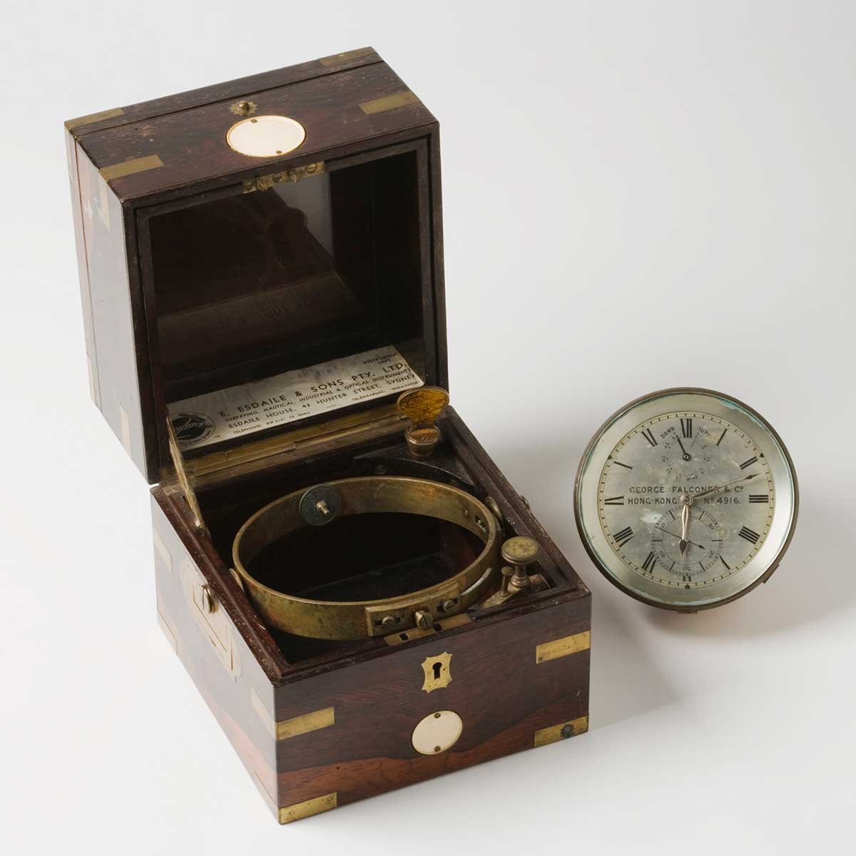 A metal chronometer sits next to a dark coloured wooden box with brass fittings. The face of the chronometer reads 'GEORGE FALCONER & Co. / HONG KONG / No. 4916.' and a label with 'E.ESDALE & SONS PTY LTD' is stuck on the inside of the box.