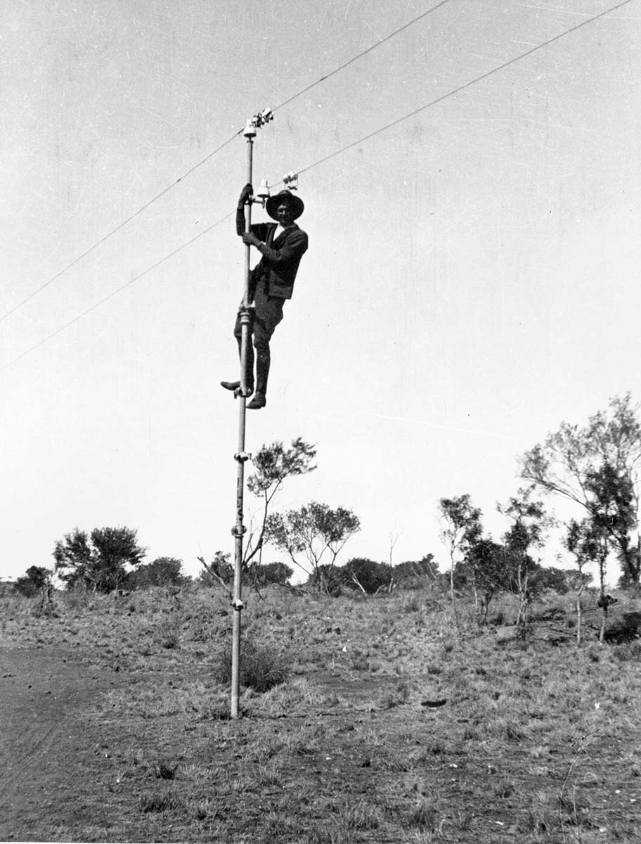 A black and white photo taken in the 1920s of a man who has climbed up to the top of a pole.