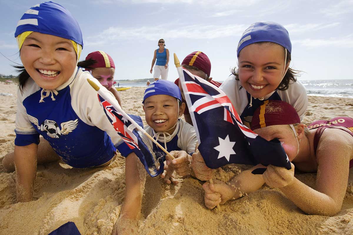 Colour photograph of six young people wearing life saving attire, laughing and smiling in the sand on a beach.