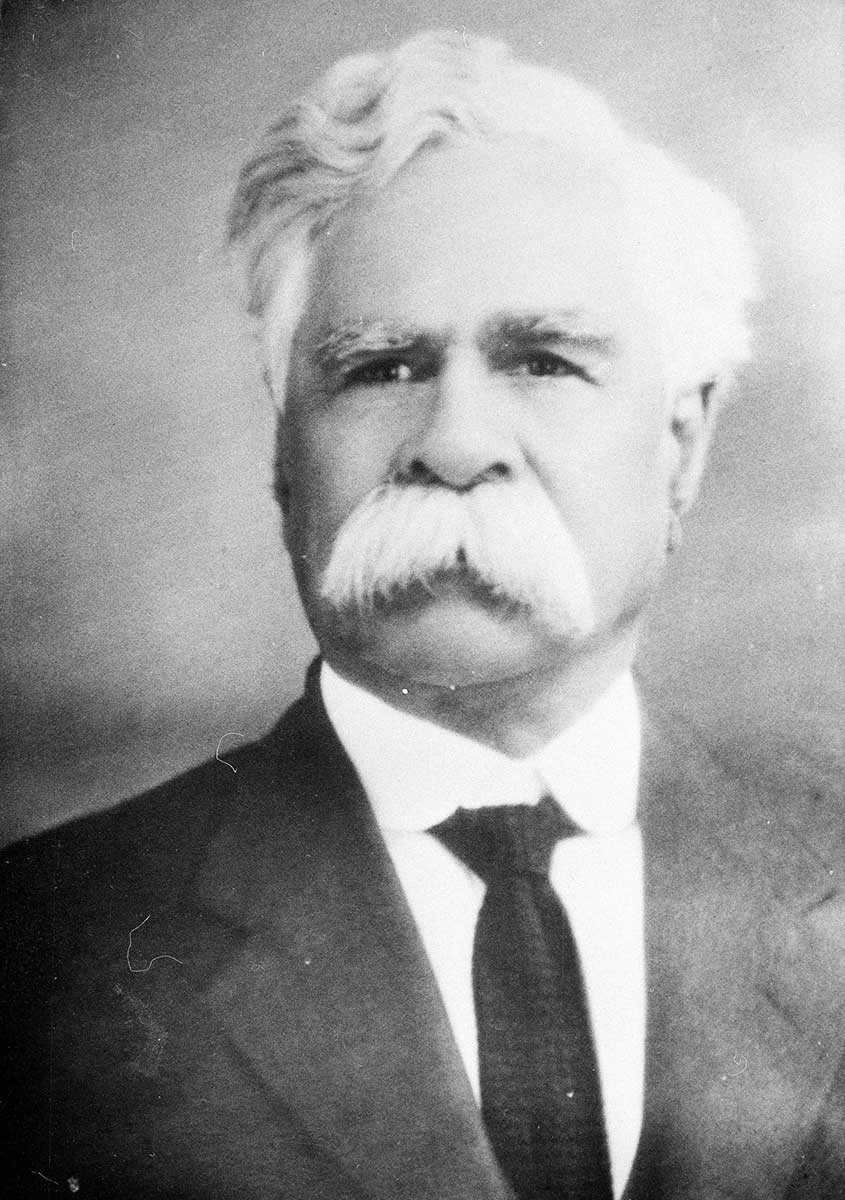 A black and white portrait photo of a white-haired man with a white moustache. 