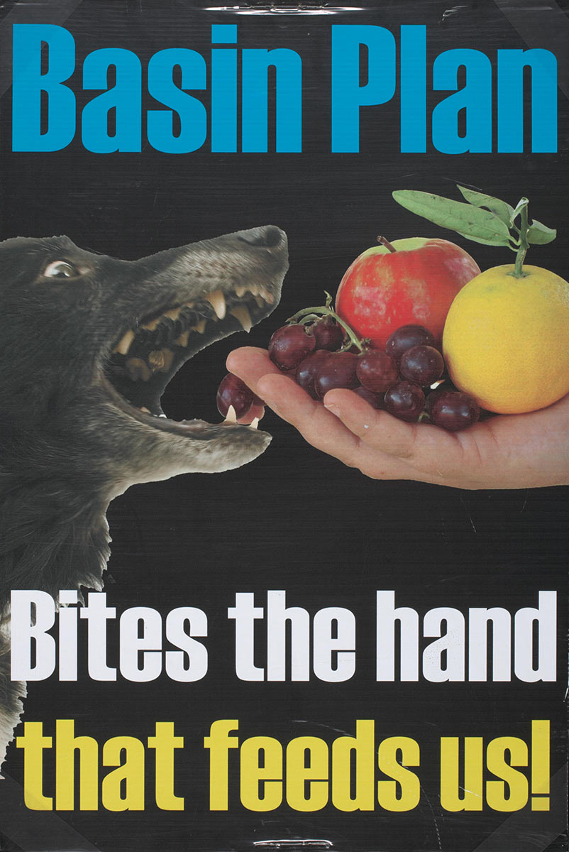 Colour photograph of a poster on a black background, with a central image of a dog biting an extended hand holding fruit. Printed text reads: 'Basin Plan. Bites the hand that feeds us!'