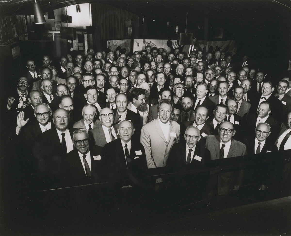Interior photo of tightly packed group of about 70 men in their 50s and 60s smiling at the camera.
