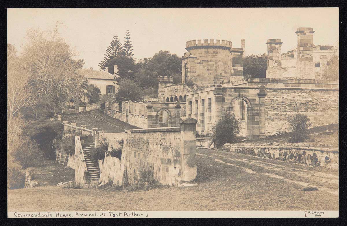 A postcard featuring a sepia photograph of a stone building with a stone wall and staircase in the foreground. Handwritten text on the front of the postcard describes the photograph as 'Commandant's House, Arsenal etc. Port Arthur]' and also '[R.C.Harvey Photo.'.