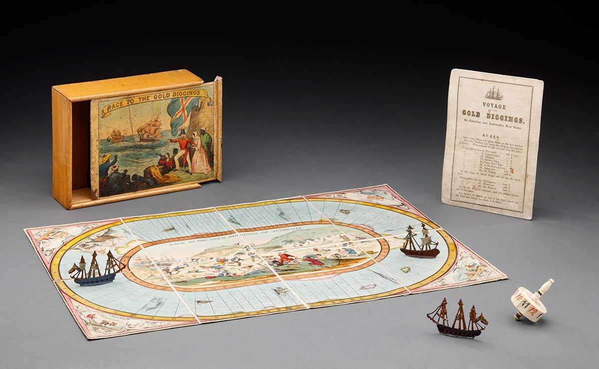 Studio photo of a game board with three pieces, an instruction manual and the box they came in. the box has a colourful illustration showing men holding a flag pointing at ships at sea. The pieces on the game board are ships and the board itself suggests that the game involves moving around an oval sea with an illustration of gold diggings in the centre.