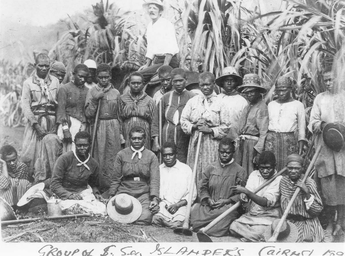 Black and white photo of a group of women in labourer clothing standing or sitting in front of a sugar cane plantation. A man on a horse behind them stares off his right.