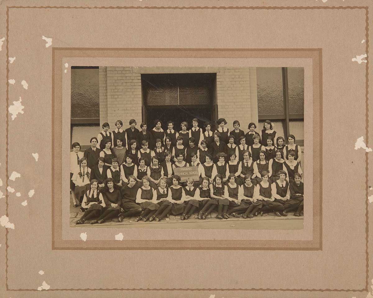 Black and white photograph depicting a large group of uniformed girls in four rows. A sign held by two girls at the centre, reads "Prahran Technical School".