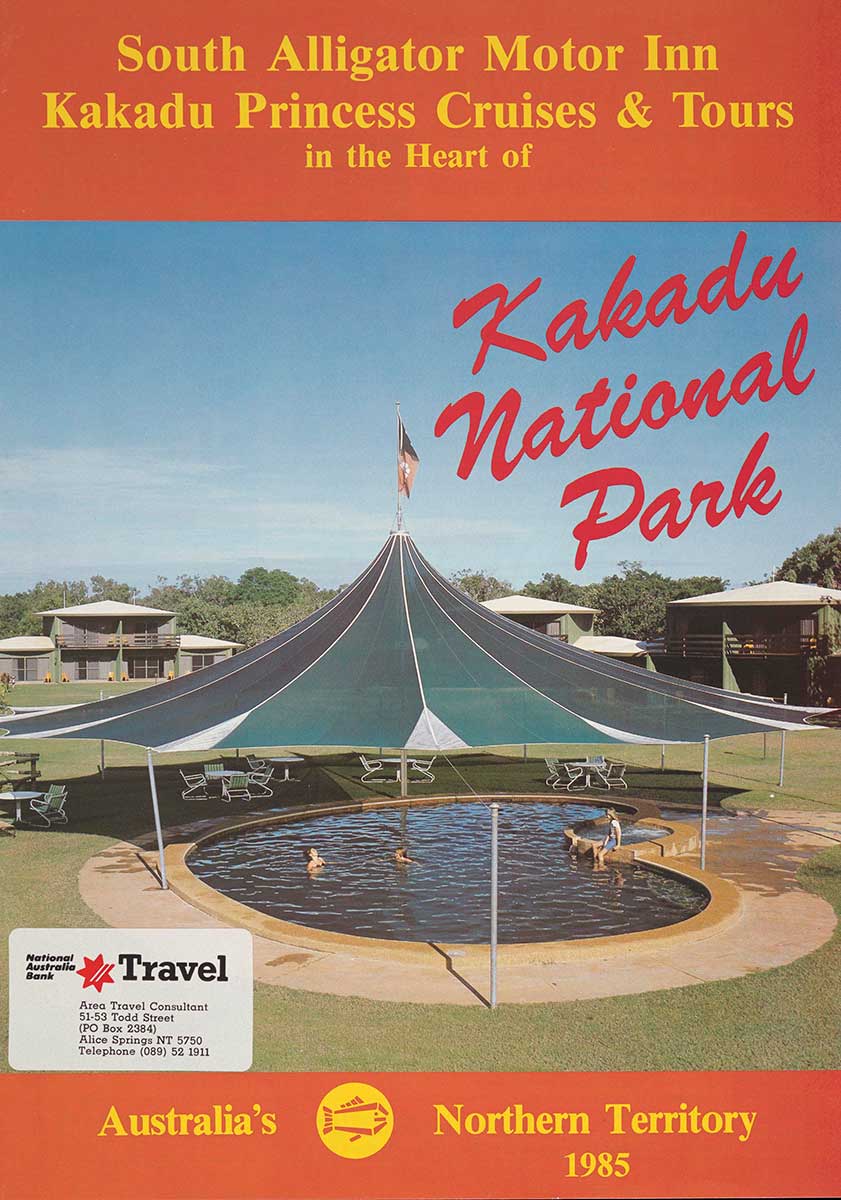 Brochure with large red inscription that reads 'Kakadu National Park.' It has an image of an apple shaped swimming pool with two people in it and a girl sitting on the edge. The swimming pool is under a tent covering with tent ends attached to metal posts. There is a red, white and black flag flying from the top of the tent. Houses surrounding the text, tables and chairs under the text.