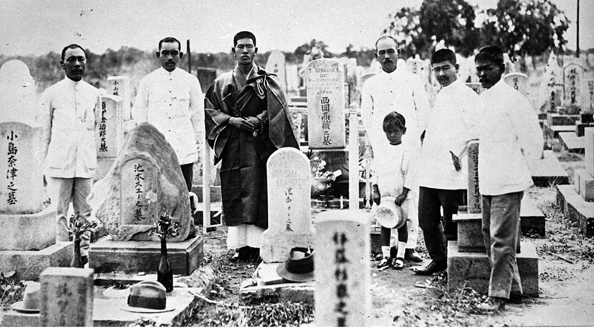 Black and white photograph of a group of men and one child standing amongst gravestones etched with Japanese text.