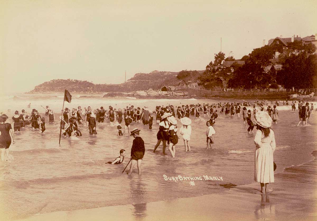 A crowded beach with well-covered bathers paddling in the shallows. A dark lifesaving flag is visible.