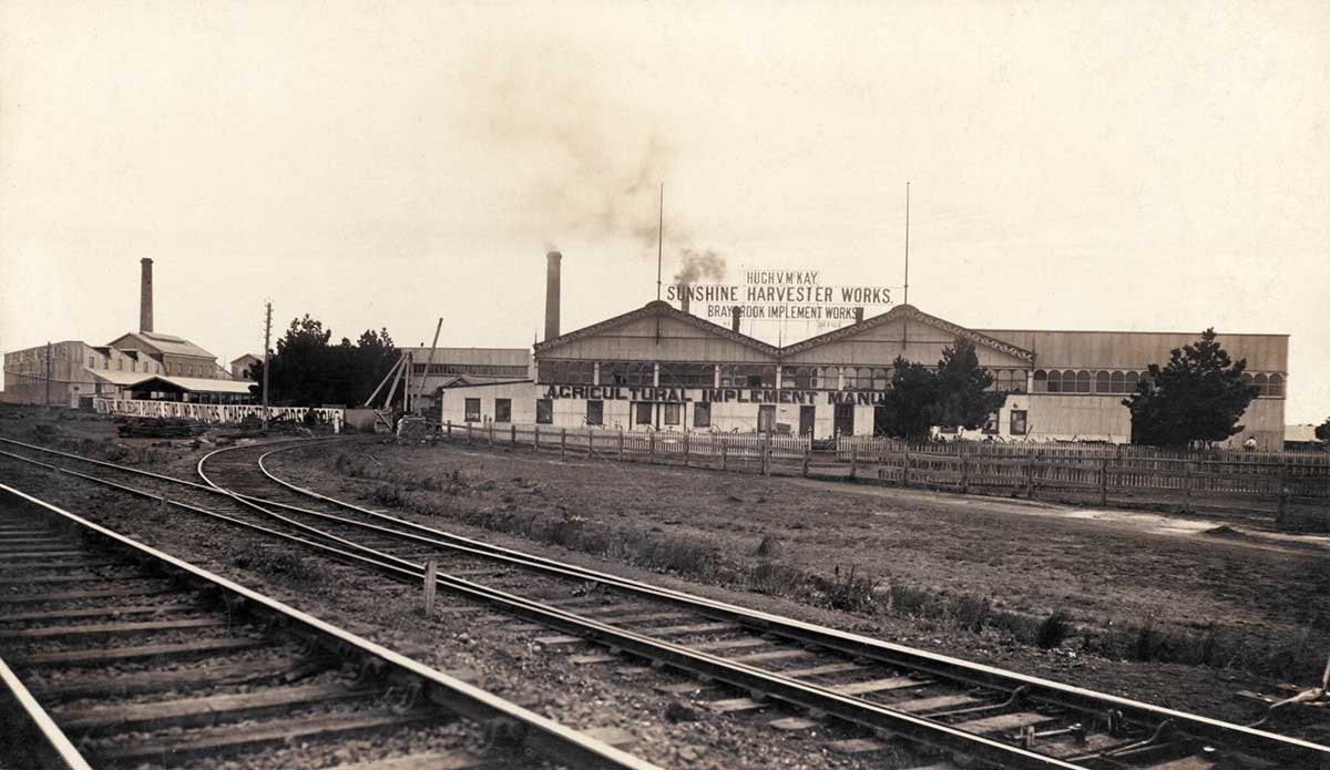 Black and white photograph of the Sunshine Harvester Works factory. Signage above one of the warehouses reads 'HUGH V McKAY / SUNSHINE HARVESTER WORKS / BRAYBROOK IMPLEMENT WORKS'. There are railway tracks in the foreground.