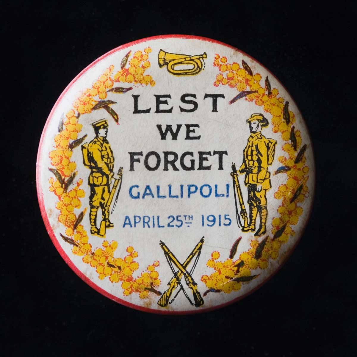 A circular metal Anzac Day badge with a plastic coating, depicting two soldiers in yellow, facing toward the centre, with a bugle at the top and crossed rifles at the bottom. They are set against a cream background with an inner border of yellow wattle blossoms, and a red outer edge. Blue and black text in the centre of the badge reads "LEST / WE / FORGET / GALLIPOLI / APRIL 25TH 1915".