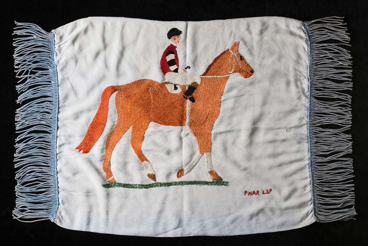 Light blue cushion cover, with blue tassels and a hand embroidered picture of a horse and jockey. 'PHAR LAP' is embroidered in the bottom right hand corner.