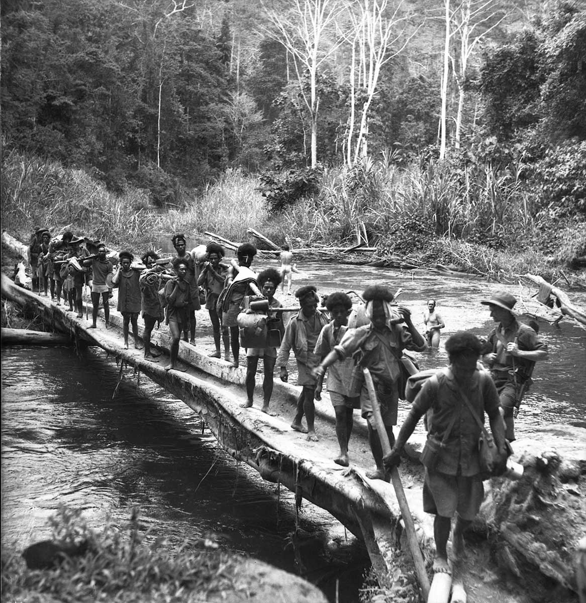 Black and white photograph of Papuan people carrying equipment across a makeshift bridge across a river. Soldiers can be seen bathing in the river.
