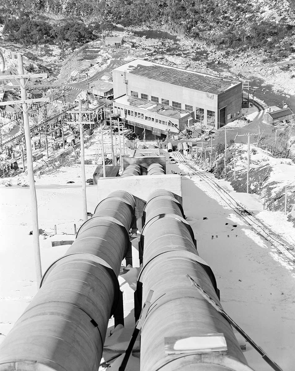 Black and white photograph of a pipeline leading to the main building of a hydro power station.