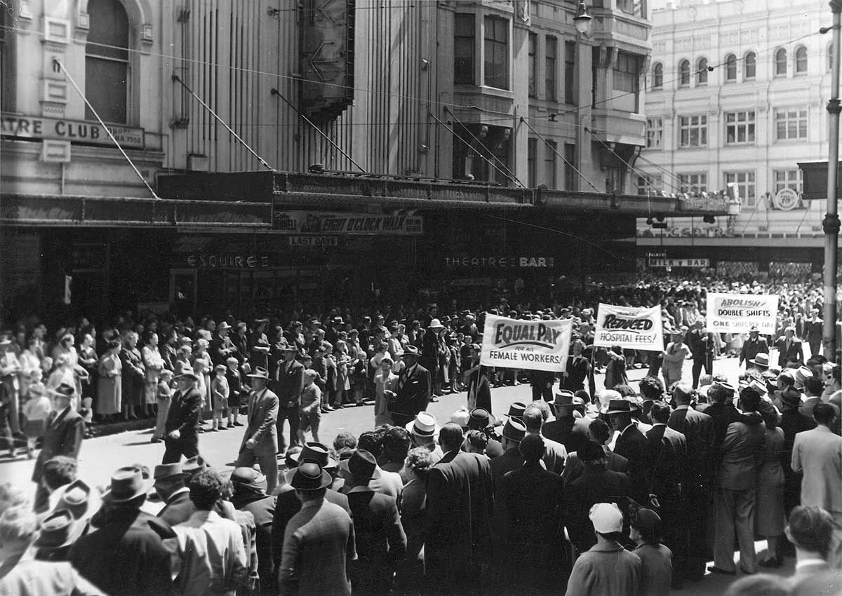 Black and white photograph of large crowds of spectators watching people walking down the street with banners supporting better working conditions.