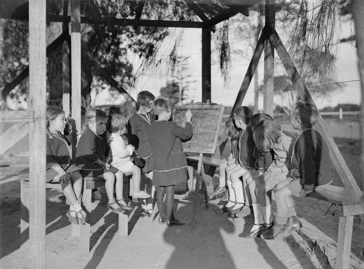 Black and white photo of a group of children around a blackboard.