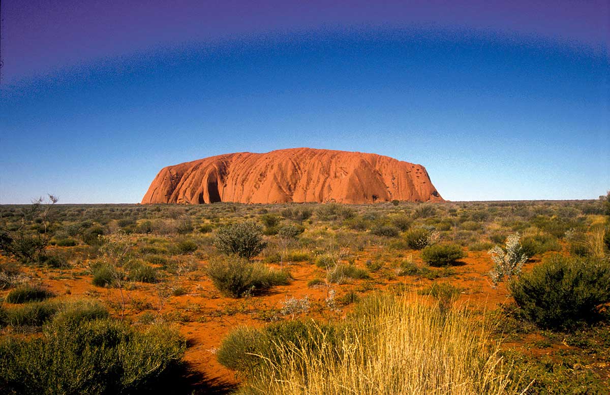 Colour photograph of an enormous rock surrounded by desert, against a clear blue sky.