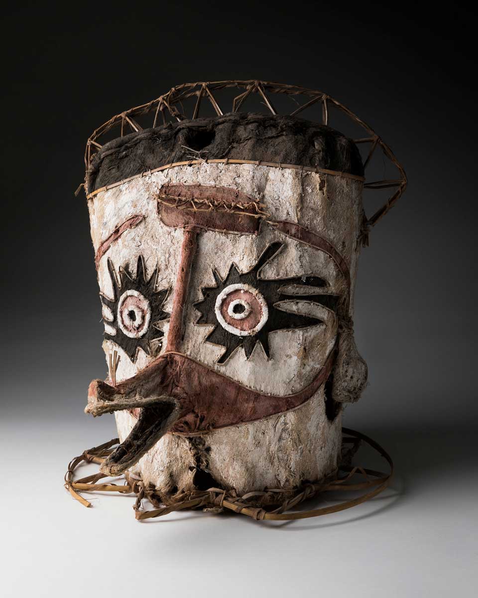A mask of tapa cloth attached to a cane frame, painted in white, black, and pink. The face has a projecting pointed mouth, and circular eyes framed by a black design. There is a cane ring around the base.