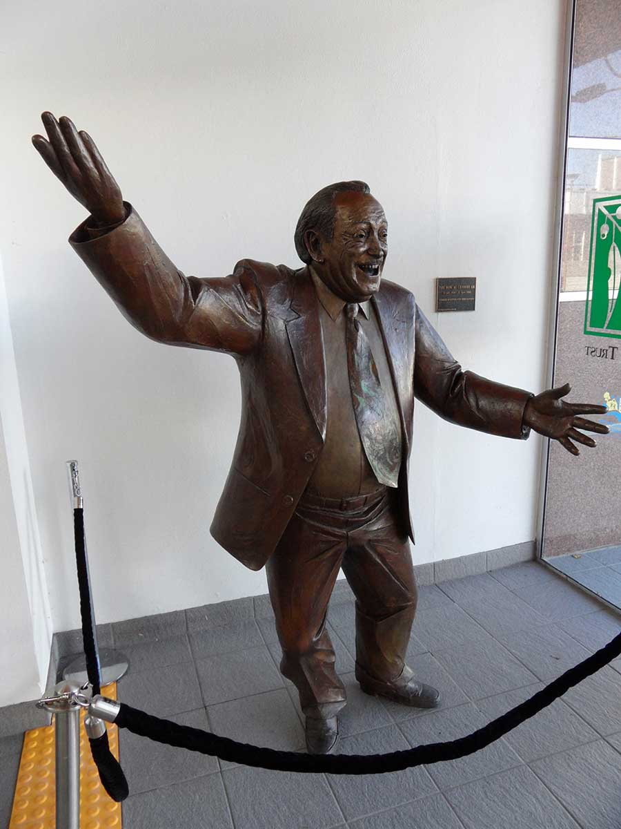 Bronze statue of a man in the entrance of a building. He is throwing his arms apart and has an amused expression.