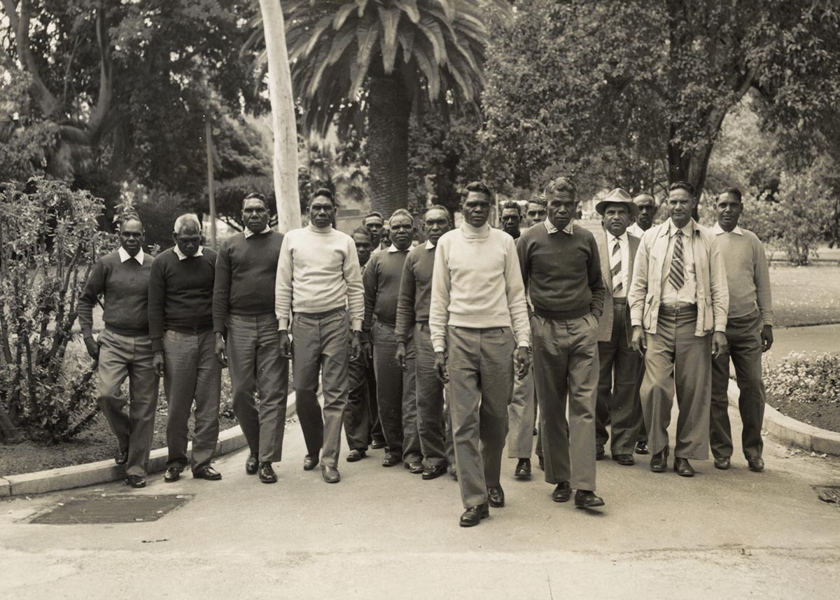 Black and white photograph of a large group of men in smart casual attire walking through a public park.