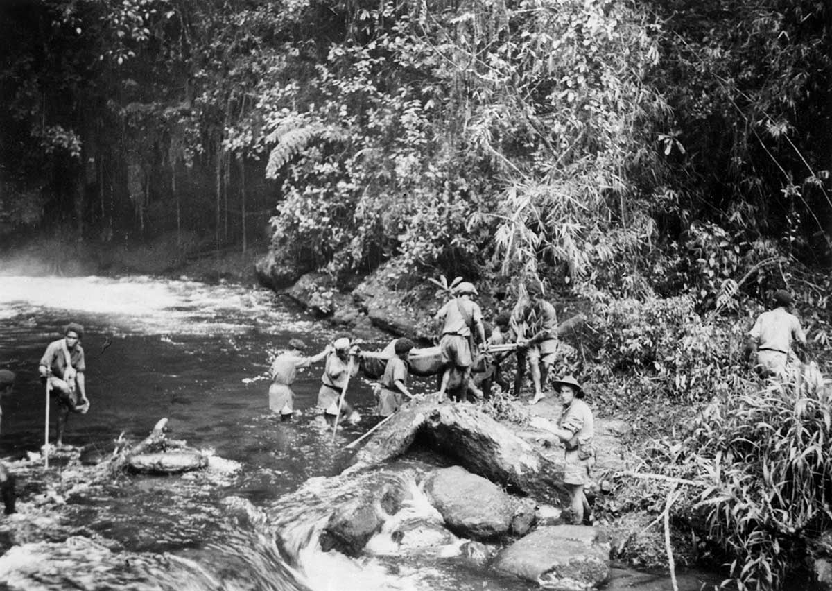 Black and white photograph of men in a jungle setting carrying a stretcher across a river.