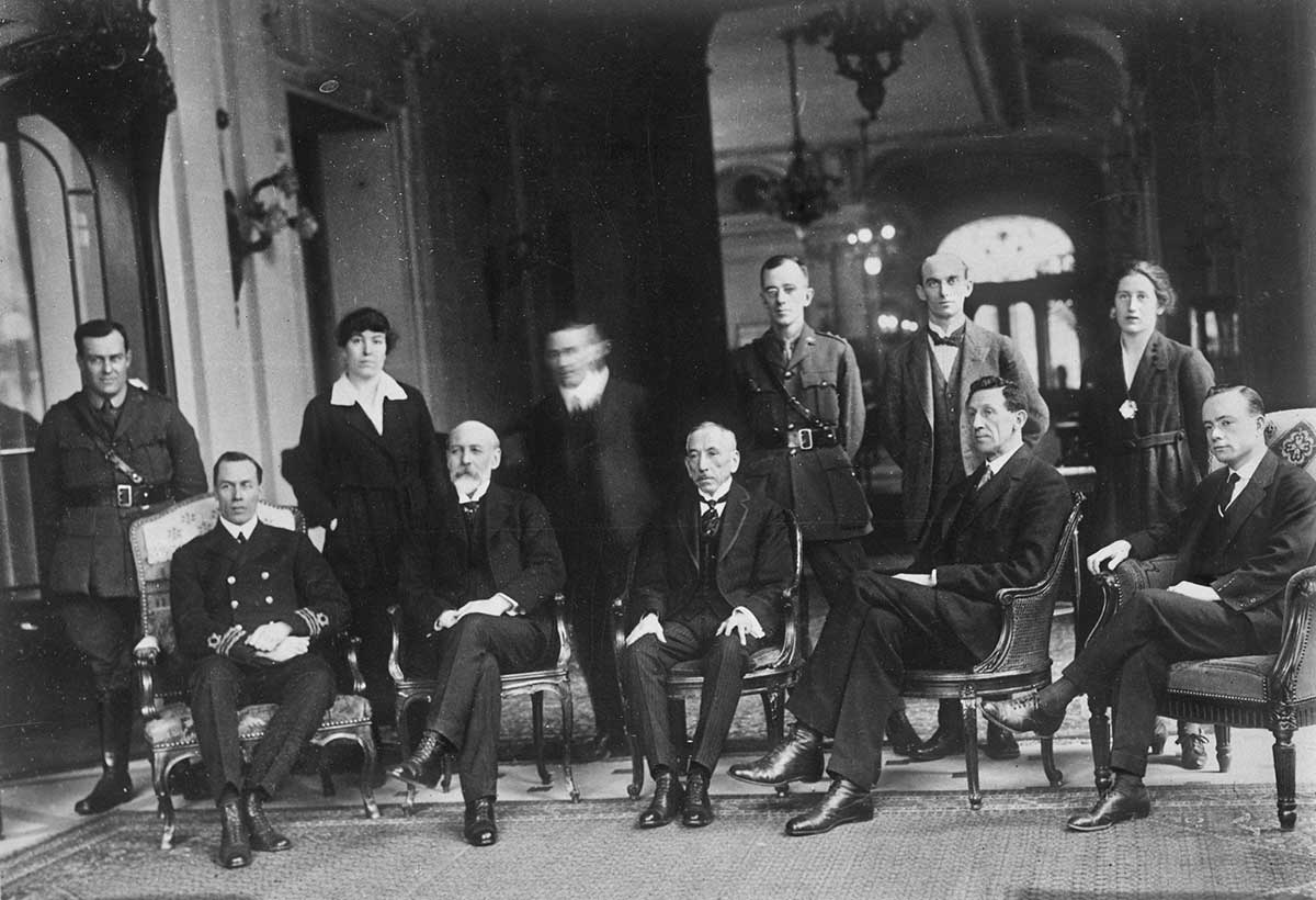 Black and white photograph of a group of seated and standing men and women in the hallway of a grand building.