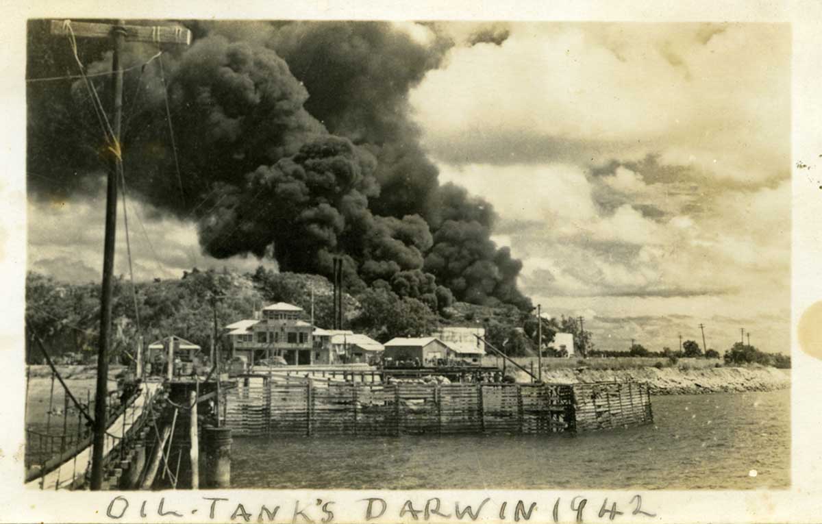 Black and white photograph of black smoke billowing from behind a hill overlooking an inlet. Hand written along the bottom is 'OIL TANK'S DARWIN 1942'.
