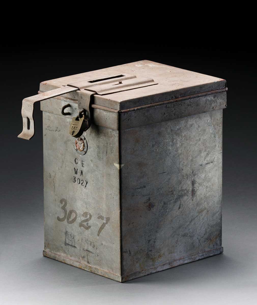 A rectangular, galvanised metal voting box with a hinged lid. The lid has two metal hasp closures, one of which has a brass padlock attached, with a key inserted. The text 'CE / WA / 3027' is inscribed on the front of the box and the number '3027' is repeated in black marker.