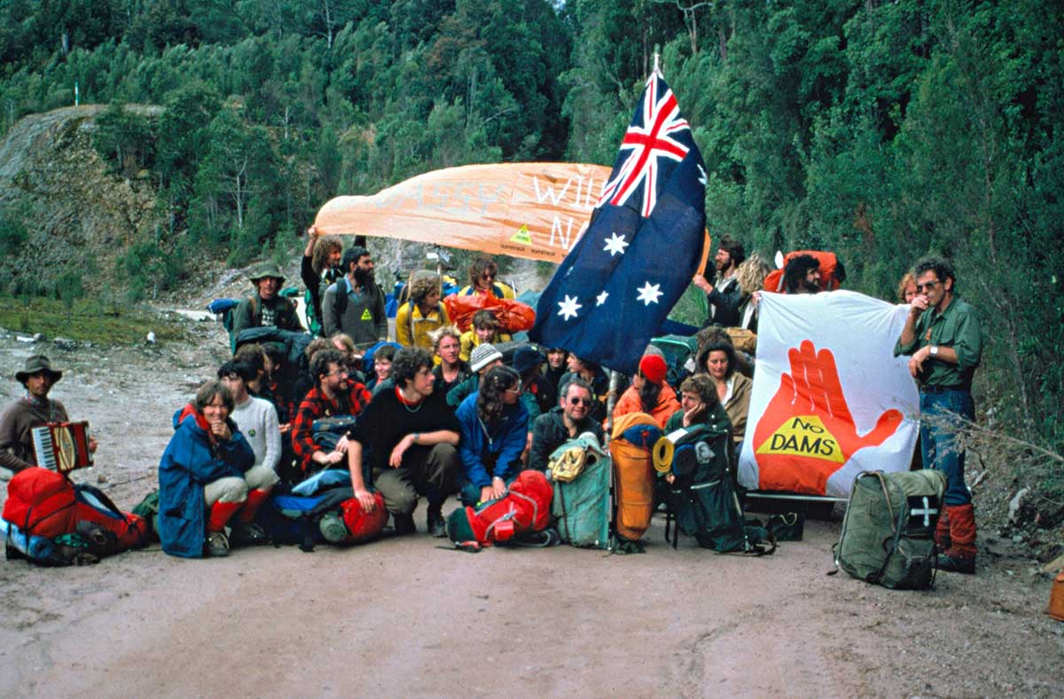 A group of protesters with hiking gear squatting or standing on a dirt road. They are holding the Australian flag and various banners, one that reads 'NO DAMS'.