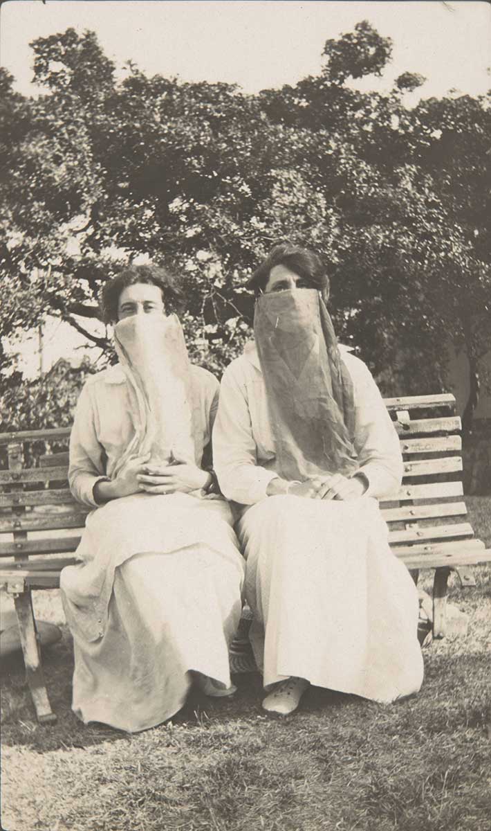 A black and white photograph of two women wearing face scarves sitting on a park bench outside.