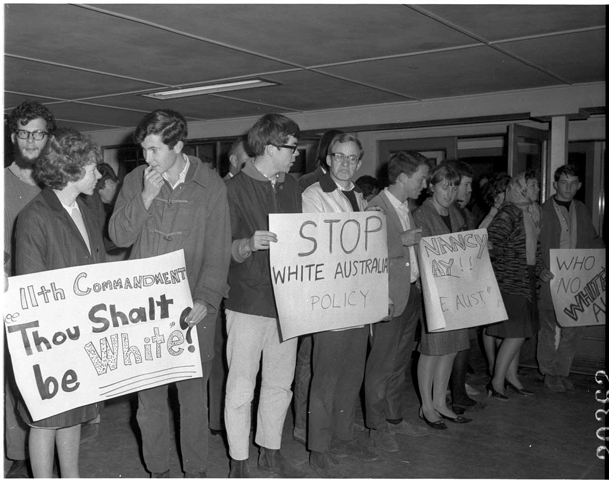 Black and white photograph of a group of men and women standing together with placards protesting against White Australia Policy.