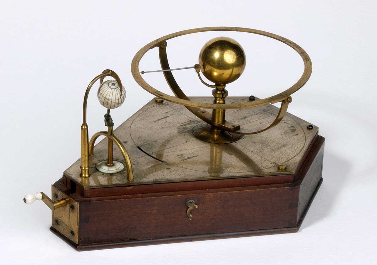 Mechanical object made from polished wood and brass fittings. It is operated by a crank fixed to one end, and globes representing Venus and Earth are mounted on top.